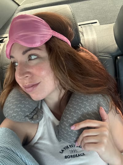The image shows a woman with long, brown hair wearing a pink sleep mask and a grey patterned neck pillow. She is seated in the back seat of a car, indicated by the black leather upholstery and a seatbelt visible. The woman is looking to her left, with a slight smile. She is wearing a white tank top with text that reads "LA VIE EST 1995 BORDEAUX". Her right hand is resting on the neck pillow. The lighting suggests it is daytime, possibly during travel. The scene does not provide specific landmarks or details to confirm a location within the city of Sychavka, Ukraine.