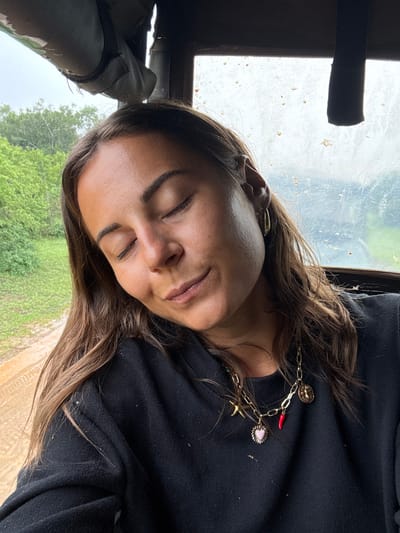 A woman with closed eyes leans against a window, her head tilted. She wears a black top and a gold necklace with various charms. Outside the window, green foliage and what appears to be a dirt path are visible. The window is slightly dirty, and a black object is visible in the upper left corner. The woman appears relaxed, possibly asleep.