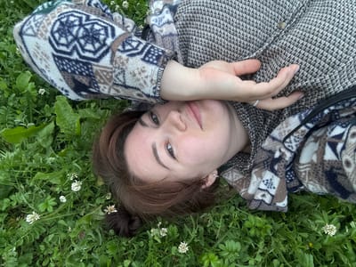 A person is lying on their back in a grassy area with small white flowers. The person has brown hair and is wearing a patterned jacket. Their right hand is raised, covering their right eye. The location is specified as Маркидоновка, Russia. The scene is outdoors.