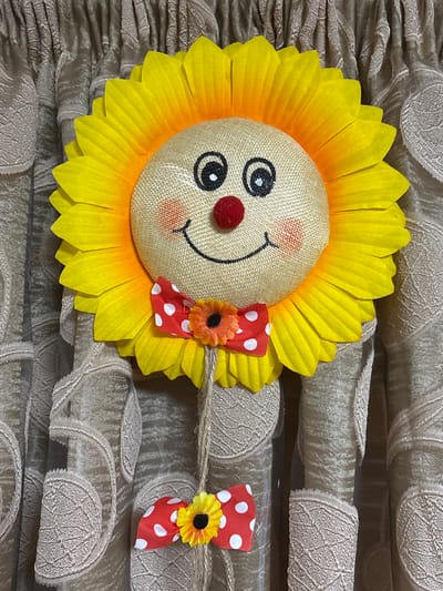 A handcrafted decorative sunflower with a smiling face is displayed against a backdrop of pleated, beige curtains with a subtle pattern. The sunflower's face is made of burlap material, featuring two black button eyes, a red pom-pom nose, rosy cheeks indicated by orange circular blush marks, and a black drawn smile. Its petals are large and vibrant yellow, transitioning to orange at the base where they connect to the face. A small red and white polka-dot bow tie with a miniature yellow and black flower is attached to the neck area of the sunflower character. Another identical bow tie with a smaller flower is suspended below the main body by a twine string. The lower part of the decoration appears to be made of two rolled-up fabric tubes, likely representing arms or legs, also adorned with the same beige patterned fabric as the background. The overall scene suggests a decorative piece placed in a domestic setting, potentially a home in Kurtovo Konare, Bulgaria.