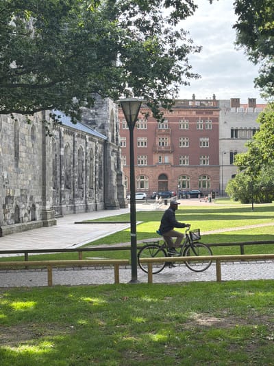 In Lund, Sweden, a person is cycling on a paved path in front of a stone building. The cyclist, wearing a dark jacket and a cap, is positioned in the middle of the frame, moving from left to right. They are riding a bicycle with a basket on the front. The stone building, which appears to be a historical structure with arched windows and intricate stonework, occupies the left side of the image. Behind the cyclist, a brick building with multiple windows stands out, and further in the background, other buildings and trees are visible. The scene is set in an outdoor environment with well-maintained green lawns and a wooden fence bordering the path. The sky is overcast with patches of blue. The lighting suggests it is daytime.