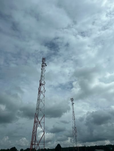 The image shows two tall telecommunication towers against a cloudy sky. The tower on the left is larger and has a red and white lattice structure, with several antennas at the top. The tower on the right is smaller, also with a lattice structure and antennas. In the distance, a line of trees and a small building are visible. The sky is filled with dark, heavy clouds, suggesting overcast weather. The overall scene is one of a rural or semi-rural landscape with essential infrastructure. This image was captured in the vicinity of Плешчаніцкі сельскі Савет, Belarus.