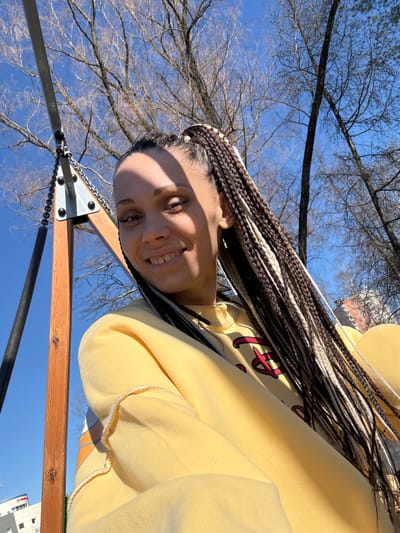 A young woman with box braids is smiling at the camera. She is wearing a yellow hoodie with a red design. The photo appears to have been taken from a low angle, looking up towards the subject and the sky. Bare tree branches are visible in the background, suggesting it may be late fall or winter. A swing set frame is visible in the bottom of the frame, partially obscuring a building. The location appears to be in an urban environment, likely Yaroslavl', Russia, based on the request.