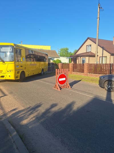 In the village of Zimkovichi, within the Zimkovichi rural council of Belarus, a bright yellow bus is parked on a paved road. To the right of the bus, a temporary wooden barrier with a red and white "do not enter" sign is placed on the road, indicating a closure. A dark-colored car is parked to the right of the barrier. The background features residential houses with brick facades and brown roofs, surrounded by wooden fences. Green trees and clear blue sky complete the scene. The lighting suggests it is daytime.