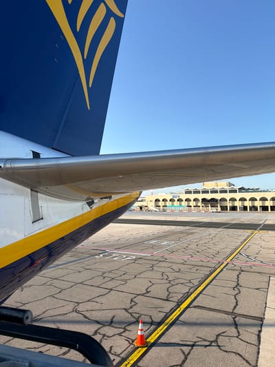 The image shows the tail and wing of a Ryanair airplane parked on the tarmac at Malta International Airport in Luqa, Malta. The aircraft's tail features the distinctive yellow harp logo against a dark blue background. The wing, with its metallic sheen, extends across the frame, partially obscuring the view of the airport terminal in the distance. The terminal building is characterized by a series of arched windows and a light-colored facade. The tarmac is marked with various lines and symbols, including a "STOP" sign painted on the ground. An orange traffic cone is visible in the foreground, placed near a yellow line on the tarmac. The sky is clear and blue, indicating fair weather. The scene depicts a typical airport environment, with the airplane ready for departure or having just arrived.