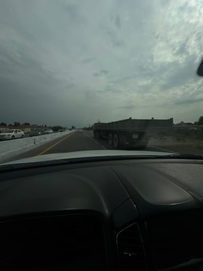 An exterior, ground-level, wide shot captures a view from inside a vehicle traveling on a highway. The sky is overcast with heavy clouds. In the foreground, the dashboard of the vehicle is visible. The highway itself features a yellow lane divider. To the left, there is a concrete barrier, and beyond it, several cars are driving or stopped. In the middle distance, a large flatbed truck is traveling in the same direction. The background shows some houses and vegetation, suggesting a semi-urban or rural environment. The location is identified as the city of Sergeli, Uzbekistan.