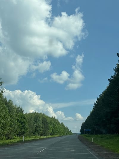The image shows a straight, paved road stretching into the distance, flanked by dense forests on either side. The sky is a bright blue, with scattered white cumulus clouds. On the right side of the road, a blue sign with white lettering is visible, with the text "ОМАРОВСКИЙ" prominently displayed. The road is marked with white lines. The environment appears to be rural or natural, possibly a highway passing through a forested region.