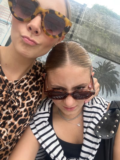 The image displays a close-up, vertical portrait of two individuals, likely females, in an outdoor public setting within an unspecified area of Ribamontán al Mar, Spain.
The individual positioned in the upper frame wears large, angular tortoise-shell sunglasses and a brown and black leopard-print top with a tie-front detail. Her mouth is pursed, suggesting a deliberate pose. Her light brown hair is pulled back.
The second individual, positioned in front and slightly to the right, has light brown hair with a distinct middle part, pulled back tightly. She wears small, oval-shaped brown tortoise-shell sunglasses lowered on her nose, looking over them with a furrowed brow and a stern expression. She wears a black and white striped top over a black tank top, accessorized with a layered silver-colored necklace featuring a small, shiny pendant. Her hands are positioned near her sunglasses, and her fingernails display a pink and white French manicure. A black bag with silver stud accents is visible on the lower right.
The background elements include a partial white sign or poster with faint red lines and text, including "TURNO 3." Behind the subjects, a grey stone wall or structure is visible, with sparse green vegetation growing on its surface. A distinct reflection of a palm tree is present in the background, indicating a nearby reflective surface or glass structure. The scene is well-lit, consistent with daylight conditions. The specific location within Ribamontán al Mar cannot be determined from the visible details.
