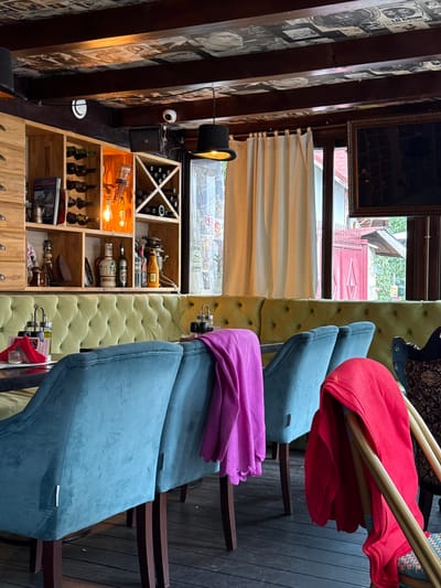 This media file depicts an indoor scene, likely a bar or restaurant, with a rustic and eclectic decor. The prominent subjects are several chairs upholstered in light green tufted fabric, some of which have a blue fabric draped over them. A red fabric is also draped over one of the chairs. To the left, a wooden shelf unit is filled with various objects. These include rows of wine bottles in the top section, decorative light bulbs and bottles in the middle sections, and what appear to be antique items like a sewing machine and an iron in the lower sections. A black top hat sits on a stand to the left of the shelves, near a security camera. The background reveals a window with a view of an outdoor area with red architecture and greenery. The overall ambiance suggests a cozy and possibly vintage setting, possibly in the evening or an interior space with artificial lighting. There is no explicit indication of an activity or event taking place, and no clear emotional cues are conveyed. The text visible on the wallpaper on the far left is fragmented and difficult to read, but appears to be in a newspaper-like style with images and headlines. The location context of Predeluț, Romania is provided.