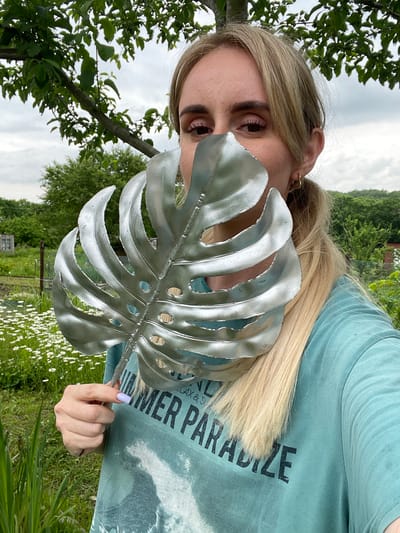 A woman with blonde hair pulled into a ponytail, wearing a teal t-shirt with the words "SUMMER PARADISE" printed on it, holds a large, silver monstera leaf in front of her face. She is outdoors in what appears to be a garden. The background shows green trees, a field of white flowers, and a cloudy sky. Her left hand, with light purple nail polish, is visible near the bottom of the leaf. The woman is looking directly at the camera, with her eyes visible above the leaf.