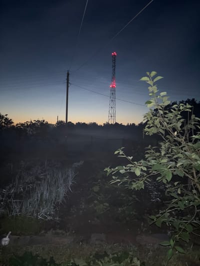 The image depicts a twilight scene with a cell tower illuminated by red lights in the distance. The cell tower is tall and metallic, with multiple levels of lights. In the foreground, there is a garden with rows of plants, possibly onions or leeks, and a small tree with green leaves on the right side of the frame. A wooden utility pole with wires is visible to the left of the cell tower. The sky is a gradient of dark blue to a lighter hue near the horizon, suggesting the transition from day to night. The overall environment appears to be rural or semi-rural, with no people visible in the frame. The location is identified as Ганчарскі сельскі Савет, Belarus.