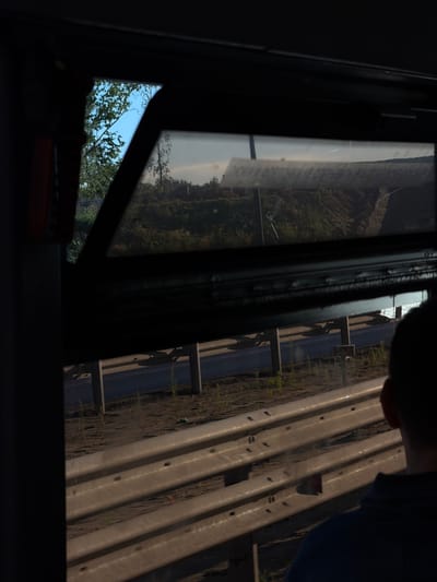 The image is taken from inside a vehicle, looking out through a partially opened window. The view is of a roadside scene, likely in the city of Дубрава-2 Снт, Russia. In the foreground, a person's head and shoulders are visible, wearing a dark blue shirt. Beyond the person and the window frame, a metal guardrail with horizontal bars is prominent. Behind the guardrail, a road or pathway is visible with some sparse vegetation. Further in the background, there are more guardrails and poles supporting what appears to be a bridge or overpass. The landscape beyond the road includes trees and a slightly elevated, rough terrain that could be a construction site or a natural embankment. The sky is visible in a section of the window, appearing light blue with some clouds. The overall impression is a transient view from a moving vehicle.