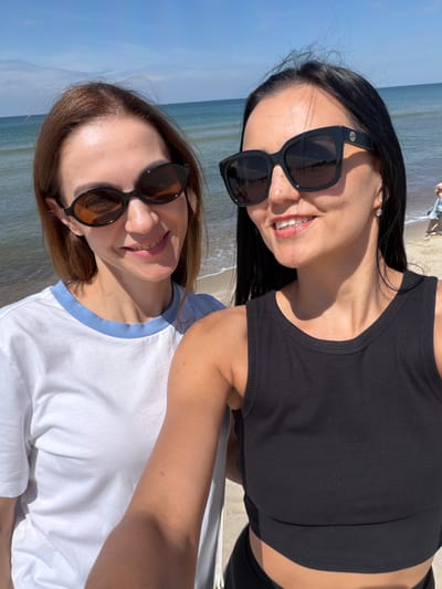 Two women are taking a selfie on a beach in Rybachy, Russia. The woman on the left has shoulder-length auburn hair and is wearing a white t-shirt with a blue trim. She is wearing dark, oval-shaped sunglasses. The woman on the right has long, dark hair and is wearing a black tank top and large, black, square-shaped sunglasses with a gold logo. She is wearing a small, diamond stud earring. They are both smiling. The background shows the sea and the sky, with a sandy beach visible to the right. The lighting suggests it is daytime.