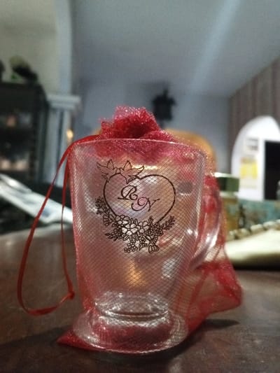 The image displays a small, clear glass, likely a votive holder or a drinking glass, encased within a sheer red mesh drawstring bag. The bag is cinched at the top, creating a ruffled closure, with a red drawstring hanging loosely to the left. A black-outlined design is present on the glass or the mesh, depicting a heart with two doves perched on its upper curve, the stylized initials "B & N" inside the heart, and a floral wreath beneath it. A subtle light source or reflection is discernible within the glass. The object rests on a dark, reflective wooden surface. The background is out of focus, revealing an indoor setting with indistinct furniture or shelving on the left, an archway or doorway on the right, and a light-colored ceiling. No individuals are present, and no actions or interactions are depicted. This scene is presented as being in Longmiang, Laos, though the image contains no specific visual identifiers for this location.
