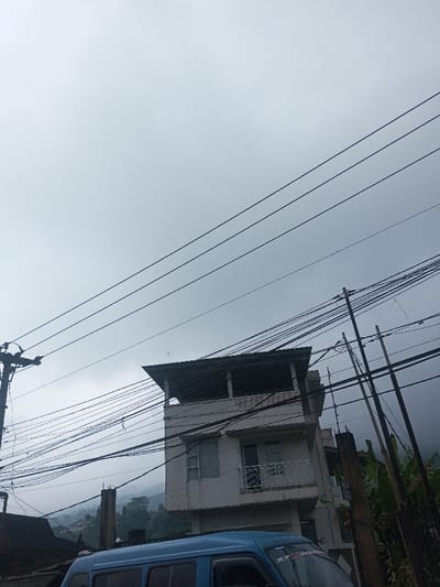 The image captures an overcast daytime street scene, dominated by a cloudy sky and a dense network of overhead utility lines. The sky occupies roughly the upper two-thirds of the frame, a uniform light grey, indicating an overcast or cloudy day with no direct sunlight.
Numerous black power lines and communication cables crisscross the upper and middle sections of the image, extending diagonally from left to right and vertically from poles. On the far left, a dark utility pole, possibly made of wood or concrete, stands, supporting many of these wires. On the right side, multiple vertical poles, appearing to be made of wood, are also heavily laden with wires, extending beyond the right edge of the frame. The sheer density of these wires is a prominent feature.
In the mid-ground, slightly to the right of the center, stands a multi-story building. It is light-colored, possibly off-white or light grey. The top visible floor features an open, covered balcony or terrace with a simple, dark roof structure overhead. Below this, the second visible floor has windows; one on the left appears partially open, while the one on the right has a white railing, suggesting another balcony. The building's architecture appears functional, with multiple sections and extensions.
In the lower part of the image, a light blue van or minivan is partially visible. Only the upper half of its side and front windshield are in view. On the windshield, there appears to be a reflective sticker or text, though its details are indecipherable due to blur and size.
The background, visible behind the building and vehicle, shows rolling hills or mountains. These distant landforms are partially obscured by what seems to be mist, fog, or low-hanging clouds, reinforcing the overcast weather impression. Green foliage, likely trees and other vegetation, covers these hills, and some faint structures can be seen on their slopes. To the immediate right of the main building, large green leaves, possibly from tropical plants like banana trees, are visible, adding to the sense of lush vegetation. To the left of the main building, a section of a dark-roofed structure is also visible.
Overall, the image presents a snapshot of a residential or semi-urban area, possibly in a hilly or mountainous region, under a dull, cloudy sky. No explicit activity is taking place, and the scene appears static.