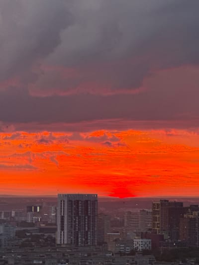 The image depicts a dramatic sunset over the city of Savenki, Russia. The sky is dominated by intense shades of orange and red, with streaks of dark grey clouds at the top. The lower portion of the sky is a vibrant, fiery orange, with the sun appearing as a bright red disc partially obscured by a cloud formation on the horizon.

In the foreground, a dense urban landscape is visible. A prominent, modern high-rise building with a distinctive facade stands out amongst other structures. Other buildings, varying in height and architectural style, fill the midground and background. Many of these buildings appear to be residential blocks, some with visible windows and architectural details. The overall impression is of a city at dusk, with the intense colors of the sunset contrasting with the dark silhouettes of the urban environment. No people or specific actions are discernible in the image.