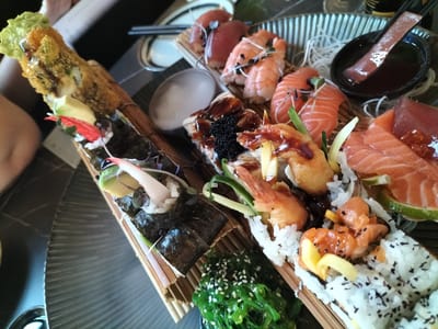 This media file showcases a close-up, overhead view of a diverse sushi platter. The main subjects are various types of sushi and sashimi, including salmon, tuna, shrimp, and what appears to be eel. Several rolls are also visible, some with a fried exterior and green avocado topping, while others are wrapped in nori with crab sticks and avocado. A small bowl of what looks like soy sauce or a dipping sauce is present, alongside a separate dish of seaweed salad sprinkled with sesame seeds. The sushi is arranged artfully on bamboo mats and a ridged serving platter.

The activity taking place is the presentation of a meal, likely a celebratory or leisurely dining experience. The lighting suggests it could be daytime, indoors, perhaps in a restaurant setting. The detailed presentation of the food indicates a focus on culinary artistry. There are no visible people or significant environmental cues to suggest the specific time of day or weather. The context provided places this scene in České Budějovice, Czechia, hinting at a dining establishment within this location. No discernible text is present in the image. The overall impression is one of a rich and appealing culinary spread.
