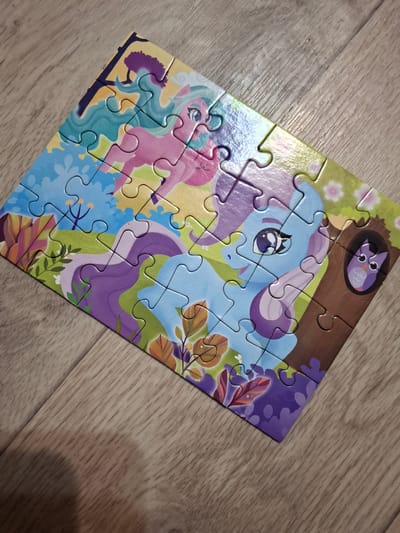 The image shows a partially completed jigsaw puzzle of a cartoon unicorn and owl, placed on a wooden surface. The puzzle is assembled except for a few unattached pieces. The color scheme of the puzzle is bright and whimsical, featuring blues, purples, pinks, and greens. The background appears to be an indoor setting with light-colored wooden flooring. The location in the city of Ozyorny, Russia is not determinable from the given image.