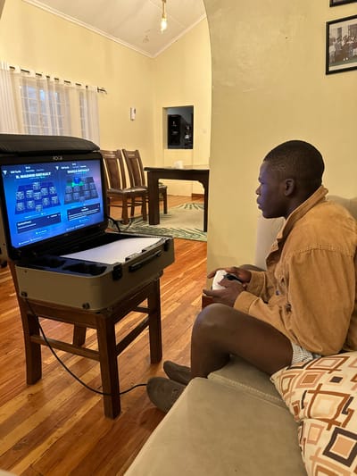 A person plays a video game on a portable gaming station sitting on a wooden stool. The gaming station's screen displays game settings. The player is seated on a couch, holding a controller. The room has cream-colored walls and wooden floors. A dining table and chairs are visible in the background. A window with sheer curtains is on the left, and a framed picture hangs on the wall above the couch.