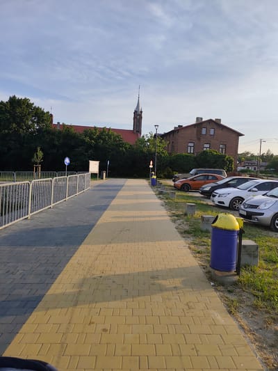 A paved walkway in Stryków, Poland, runs alongside a grassy area where several cars are parked. The walkway is made of two different colored paved sections, one gray and one yellow, with a distinct shadow line separating them. A metal fence runs along the left side of the walkway, separating it from a gravelly area. In the background, a brick building with a red roof and a tall church spire are visible amongst trees. The sky is blue with some wispy clouds. A blue trash can with a yellow lid is positioned near the walkway. The image appears to have been taken on a sunny day, likely in the late afternoon or early evening due to the long shadows. There are no people visible in the image.
