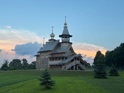 An exterior view of a traditional wooden Orthodox church with multiple onion domes and crosses is captured under a twilight sky. The church, constructed from logs, features intricate wooden detailing, including a covered walkway and a prominent staircase leading to an elevated entrance. The building is situated on a grassy hill, with several young evergreen trees in the foreground. In the background, a line of mature trees forms a dense forest, and a faint sunset can be seen through the clouds. The sky is a gradient of blue, transitioning to pale orange and pink hues near the horizon. There are no people visible in the scene. The location is identified as the village of Neman, in the Neman Rural Council, Belarus.