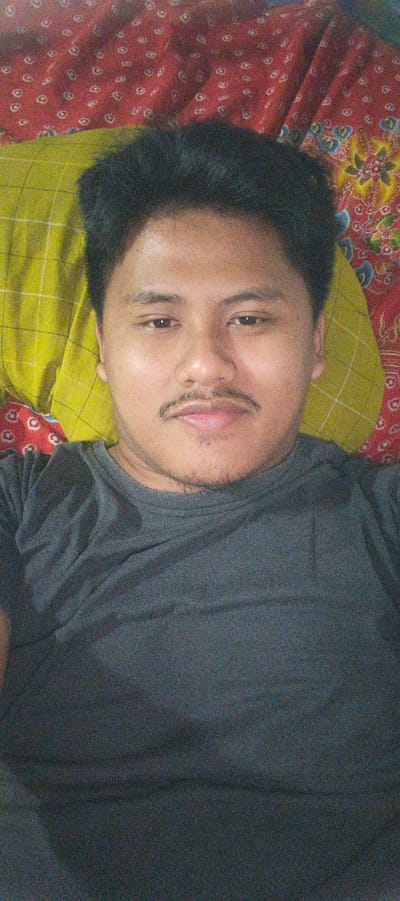 A close-up, head-and-shoulders selfie is shown, taken from a slightly elevated angle. The subject is a male with dark hair and a mustache, wearing a dark gray t-shirt. He is lying down on a bed with a yellow patterned pillow behind his head, and a red patterned blanket under the pillow. The image appears to be taken indoors in Kemuning, Indonesia.