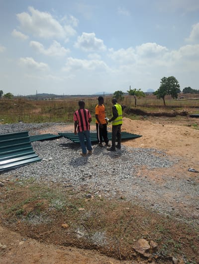 This outdoor image, rotated 90 degrees to the left, captures three men on a development site in Karsana 2, Nigeria, under a bright, cloudy sky. The ground consists of reddish-brown earth, gravel, and sparse dry vegetation.
Three men, all Black, are the primary human subjects. One man wears a bright yellow safety vest over a dark long-sleeved shirt and dark pants, standing near a pile of green metal sheets. Another man, standing beside him, is dressed in an orange t-shirt with the word "EMIRATES" clearly visible on the front, and dark pants. Both appear to be observing or discussing something. A third man, further back, wears a red and black striped t-shirt and blue jeans, standing next to a longer row of stacked green metal sheets, possibly corrugated roofing panels.
In the foreground and midground, several stacks of new, dark green metal sheets are laid out on the gravel and dirt. A small pile of light brown sand is also visible. The immediate surroundings show patches of gravel and dirt, with some scattered rocks and dry weeds. In the background, there's an expansive landscape with distant trees, low hills, and what appears to be undeveloped land stretching to the horizon.
The activity appears to be related to construction or site preparation, with the men possibly inspecting materials or planning work. The time of day is clearly daylight, likely mid-morning or afternoon, given the bright illumination and distinct shadows. The weather is fair with a blue sky dotted with scattered white cumulus clouds. The general atmosphere suggests a work-in-progress environment.
