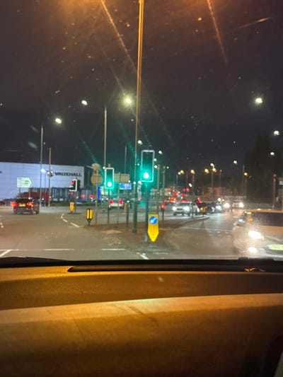 The image, captured at night from inside a vehicle, depicts a view of the intersection of Manchester Road and Rochdale Road in Bury, United Kingdom. The lower portion of the image shows the illuminated dashboard and windshield of the capturing vehicle.
Two primary traffic signals in the center-right are illuminated green, indicating permission to proceed. A third traffic signal, located further to the left, displays a red light. The road surface in the foreground and mid-ground appears reflective, suggesting damp conditions.
To the left, a large, white commercial building bearing the "VAUXHALL" logo is clearly visible. Multiple vehicles are present on the road: a dark car with red taillights is positioned on the far left, while several other vehicles with illuminated headlights are visible approaching the intersection from various directions in the mid-ground and background.
Road infrastructure includes multiple streetlights, road markings, and small traffic islands with yellow bollards. A large white directional sign with a green arrow pointing left is partially visible near the Vauxhall dealership. Other yellow warning signs and blue circular regulatory signs are also present throughout the intersection. No pedestrians are visibly discernible. The sky is dark, consistent with nighttime conditions.