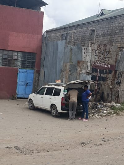 This image depicts an outdoor scene in Matungulu west ward, Kenya, under an overcast sky. A white, five-door station wagon, identified as a taxi by a roof sign, is parked on unpaved ground with its rear hatch open. Two individuals are standing at the rear of the vehicle. A male in a light brown t-shirt and grey trousers is positioned facing into the open trunk, engaged with its contents. A female in a blue t-shirt, blue jeans, and pink slip-on sandals stands beside him, observing the trunk.
To the left of the vehicle, a red-painted building features multiple grid-patterned windows and a closed blue double door. Faint white text reading "SERVICES OFFERED" is visible on its upper wall. To the right of the vehicle, an unpaved area contains a pile of discarded rubbish. This area is bordered by an irregular wall constructed from various corrugated metal sheets, some severely rusted. Written in black paint on one of these sheets is "YARD TO LET" followed by the phone number "0724 314469." Behind the corrugated metal barrier, a two-story building constructed from grey concrete blocks with a green sloped roof is visible, featuring several window openings, some of which are barred. The ground throughout the visible area is dirt and gravel.