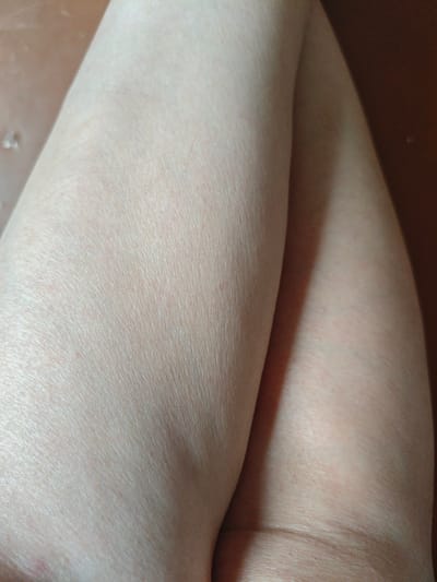 The image is a close-up, low-angle shot of a person's bare legs, positioned in a crossed pose. The skin appears pale and is illuminated by soft lighting, highlighting the texture and fine hairs. One leg is positioned in front of the other, creating a gentle curve where they meet. The background is a blurred, warm brown surface, possibly a floor or furniture. There are no identifiable landmarks or elements that would suggest a specific location within Sumgayit, Azerbaijan. The focus is entirely on the details of the skin and the form of the legs.