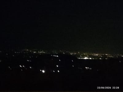 This is a nighttime, landscape photograph taken from an elevated position overlooking a city or town. The foreground is dark and indistinct, suggesting it is a natural landscape such as hills or fields. In the midground and background, numerous lights are visible, indicating urban development. These lights appear as blurred streaks and dots, characteristic of a long exposure or the limitations of low-light photography. The lights form a horizon line across the image, suggesting a settlement spread out below. Some clusters of lights appear more concentrated, possibly representing the denser areas of a town. The overall scene depicts a view of human habitation at night.
A timestamp is visible in the lower right corner of the image, reading "23/06/2026 22:28". This indicates the photograph was taken on June 23, 2026, at 10:28 PM. The time of day is clearly night, and the dark sky supports this. There are no discernible weather conditions, but the darkness suggests a clear or at least not overcast night. No people or specific objects are clearly identifiable. The location context of Scordia, Italy, would suggest this is a view of that town or its surroundings. The mood is quiet and serene, evoked by the darkness punctuated by the distant glow of lights.