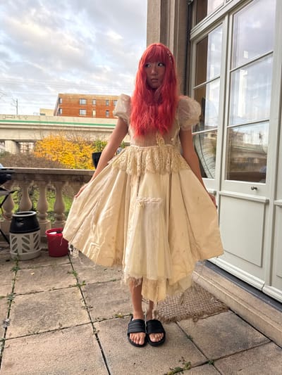 A single female individual with long, straight, bright red hair, a side fringe, and a small hair clip on the right side stands centrally on a stone-paved patio in Wallisellen, Switzerland. She faces slightly left of the camera with a neutral expression, wearing a voluminous, distressed, light cream-colored dress with puffed sheer sleeves, a lace-detailed bodice, and a multi-layered skirt displaying frayed edges and minor patches. Black slide sandals with white stripes are on her feet. Her hands are positioned on the sides of her skirt.
To her immediate right is a light-colored building facade featuring a large, multi-paned double door with a visible handle, a fluted column, and a coir doormat. Reflections on the door's glass include interior elements and the reversed German word "HUUSMEISTER". To her left, a stone balustrade separates the patio from a lower area with prominent vibrant yellow autumn foliage. In the midground, an elevated concrete bridge with overhead power lines extends horizontally, with a reddish-brown building visible beyond it. The sky is overcast with scattered clouds. On the patio to the left of the individual, a dark grey bin with a black lid, a white decorative planter, and a red bucket are present.