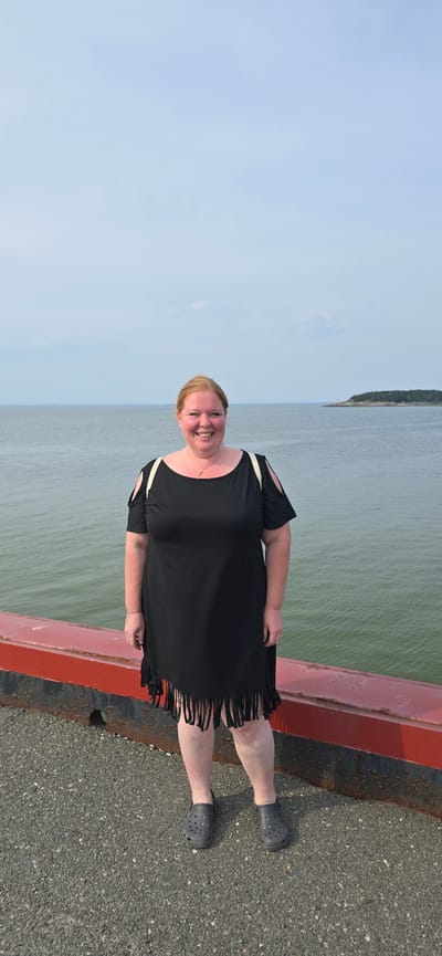 A medium shot captures a woman with blonde hair, wearing a black fringed dress and dark gray Crocs, standing on a gravel surface next to a red railing. The woman is smiling and facing the camera. Behind her, the ocean stretches out under a pale blue sky. In the distance, a small, tree-covered island is visible. The scene appears to be a waterfront location in Trois-Pistoles, Canada.