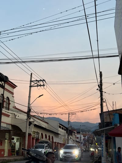 The image captures a street scene in Timaná, Colombia, at dusk or dawn, looking down a road with active traffic. The sky transitions from a light blue overhead to hues of pink and orange towards the horizon, with two distinct bright celestial objects visible in the upper left quadrant. A dense network of black electrical and communication wires crisscrosses the sky, supported by numerous concrete utility poles. Birds are perched along several of these wires.

On the left side of the street, multi-story buildings with white facades and red trim feature arched windows with dark railings. Storefronts are visible at ground level, one displaying "Carnes" and "Acai." On the right, buildings constructed with exposed brick and concrete elements are present, with a partial vertical sign for "BILLARÉS" visible on the far right. Distant mountains form a dark silhouette against the horizon.

The asphalt road appears wet in areas, reflecting light. Vehicular traffic is in motion. In the immediate foreground left, the head and shoulder of a male individual are visible, possibly on a dark motorcycle. A white pickup truck with a green and white logo on its door and roof racks is situated mid-left. A white SUV (Kia Seltos, license plate GTK-9429) with illuminated headlights is prominently centered, facing the viewer. Further down the street, additional vehicles, including motorcycles, are visible with their headlights illuminated.
