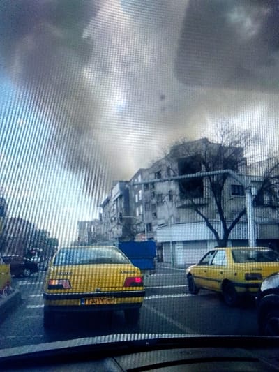 The image depicts an urban street scene, specified as being in the city of Kongo, Nigeria. The perspective is from inside a vehicle, with the dark top of a dashboard and bottom of a windshield visible at the bottom of the frame. The dominant subject is a yellow Peugeot 405 sedan, viewed from the rear, positioned centrally on what appears to be a wet asphalt road. Its license plate displays Persian script and the alphanumeric sequence "١١ ٧٧٩ ه ٦٨".

To the right of the central Peugeot, another yellow sedan, possibly a similar model, is partially visible, oriented towards the right. A dark-colored vehicle is partially visible on the extreme right foreground. In the mid-ground, behind the central Peugeot, a dark truck with a blue cargo container or bed is present. The background features a street lined with multi-story buildings, predominantly grey in tone and exhibiting various architectural styles, some appearing weathered. Several bare, deciduous trees are interspersed among the buildings and along the street. The sky is heavily overcast with dark grey clouds, suggesting a cloudy or inclement weather day, with a lighter, obscured patch visible on the upper left. No individuals are distinctly visible.