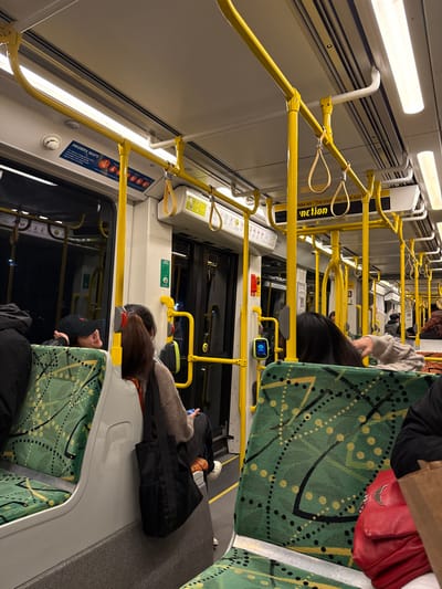 The image shows the interior of a train carriage. The carriage is brightly lit with white overhead fluorescent lights. The walls of the carriage are white with yellow handrails and yellow support beams. The carriage is sparsely populated, with only a few passengers seated on the green-and-black patterned seats. The passengers are all wearing dark clothing. The carriage is clean and appears to be well-maintained. There is a sign on the wall to the right of the image that reads "Hold on. The tram may stop suddenly". The sign is yellow and has a black background. The image is taken from inside the carriage, looking towards the front of the train. The image is slightly out of focus and blurry. The image was taken on a train in Melbourne, Australia, at the coordinates -37.829, 144.955, which corresponds to the Southbank area of Melbourne.
