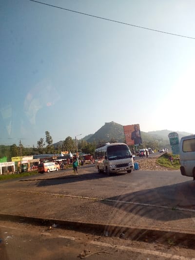 A bustling scene unfolds at a roadside market and intersection. A tan mini-bus sits prominently in the center, with a person standing nearby. Other vehicles, including a colorful van, populate the street. Shops line the side of the road, and a large billboard towers above, displaying a figure. A prominent mountain rises in the background under a clear sky. The image's perspective suggests it was taken from inside another vehicle.