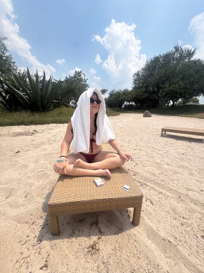 A woman wearing a dark-colored bikini and sunglasses is sitting cross-legged on a wicker-style lounger on a sandy beach. She has a white towel draped over her head. In her right hand, she holds a lit cigarette. On the lounger near her legs are a pack of cigarettes and a lighter. The lounger sits on sand, which transitions into grassy areas with various plants and trees in the background. The sky is blue with scattered white clouds. The location is Pueblo Nuevo de Jasso, Mexico.