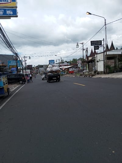 The image displays a wide-angle street scene in the city of Taohom, Laos, under an overcast sky. A dark asphalt road dominates the lower frame, featuring a faded yellow dashed center line. On the far left, a blue and yellow food cart is partially visible at the curb.
On the left side of the road, a dark-colored SUV is parked, and approximately four individuals are visible on the sidewalk. Two females are prominent, both wearing head coverings, one in a pink top and the other in a dark top, appearing to walk or stand. Behind them are commercial buildings, one bearing a large sign in Indonesian that reads "AYAM GORENG" (Fried Chicken) and "ANGKRINGAN." A dense tangle of overhead wires crisscrosses the left side of the frame above these buildings.
In the mid-ground, a black pickup truck, heavily overloaded with large white sacks, proceeds away from the viewer on the right side of the road. Its license plate, partially visible, begins with "P 12XX YX". Behind it, a white SUV is also present.
On the right side of the street, a series of buildings feature various commercial signs. A prominent black sign, supported by a street lamp post, displays "BUBUR AYAM NASI PAKET" (Chicken Porridge Rice Packet) in Indonesian. Adjacent to this, a building with distinctive traditional-style roof ornamentation, resembling Balinese or Indonesian architecture, is visible. A sign on this building reads "HOTEL AKAN MAKAN" (Hotel Will Eat) and "MINUM" (Drink) in Indonesian. Streetlights are intermittently spaced along the street.
The background shows the road extending into the distance, flanked by more businesses and billboards, with hills visible under the cloudy horizon.