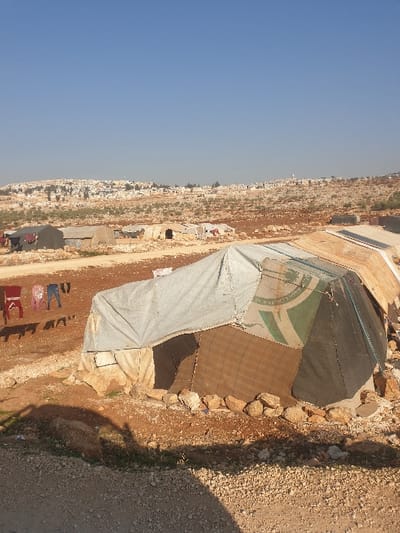 The image captures a rudimentary settlement nestled in a dry, rocky, and hilly landscape under a clear blue sky, illuminated by bright daylight. No people are visible.
In the foreground and midground, several makeshift shelters dominate the scene. The most prominent is a large, tent-like structure constructed from various materials, including dark green and lighter-colored tarpaulins. On one section of the dark green fabric, there is a distinct circular green logo featuring a white emblem resembling a stylized tree or plant, accompanied by white Arabic text that is not fully legible but appears to be part of an organizational name. Attached to this main tent, or nearby, is another shelter made of lighter-colored tarpaulin.
In front of the lighter-colored tent, several items of clothing are laid out on the bare ground, suggesting they are drying, indicating human habitation. Two simple red chairs are also visible near the drying clothes. A rough dirt track or path traverses the scene, with piles of rocks forming low walls or boundaries along its edges and around the shelters.
The background reveals a rugged, rising terrain covered with sparse vegetation and scattered buildings, suggesting a distant village or town. The overall scene depicts an informal or temporary settlement in an arid environment, consistent with the provided context of Killi, Syria.