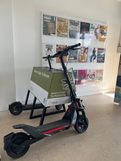 The scene depicts an interior view within a public or commercial building. While the prompt specifies the location as the city of La Unión, Spain, prominent text on the electric scooter charging station and several posters clearly indicates "Santa Lucía de Tirajana."
In the foreground, two electric scooters are parked. The primary scooter, positioned centrally and closest to the viewer, is black and red with robust features, including visible disc brakes and thick tires. It is partially obscured by a dark grey handle and front wheel mechanism. Behind it, to the left, is a second, mostly black electric scooter. Both scooters are positioned in front of a dedicated charging and parking station.
The charging station features a dark metallic frame supporting a rectangular box-like structure, predominantly colored in a two-tone scheme of olive green and white. Text on the station, visible in Spanish, reads "Aparca y Carga Tu Patinete" (Park and Charge Your Scooter) and includes the logo for "Santa Lucía de Tirajana."
The background consists of a plain white wall displaying a grid of approximately sixteen to eighteen informational and cultural event posters. These posters, enclosed in clear plastic sleeves, advertise various activities and services, including "Taller de Poesía" (Poetry Workshop), "Biblioteca Central" (Central Library) with operational hours, "Tarzán," "Curso de Photoshop II" (Photoshop II Course), "Ciudad Limpia" (Clean City), and "Susa Caramelo." Several posters also display the "Santa Lucía de Tirajana" branding.
The floor is a light, possibly beige, tiled surface. The area is well-lit, with a visible light source from the right, casting soft shadows. No individuals are present in the image.