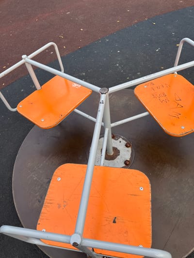 A top-down, close-up shot shows an orange merry-go-round on a playground in Verkhnerussky, Russia. The merry-go-round has three orange seats, each with a grey metal bar for support. The seats appear worn with scratches and some graffiti. The ground around the merry-go-round is a mix of dark grey and reddish-brown rubberized playground surfacing, with a few fallen leaves visible. The merry-go-round itself is made of grey metal.