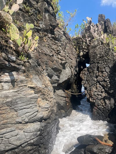 A natural rock formation with a gap carved by the sea is featured in this image, set against a bright blue sky with scattered clouds. The rough, dark gray rock face is covered in patches of green prickly pear cactus. Waves crash and churn in the narrow passage, creating white foam as they surge through. A thick rope is strung horizontally across the lower part of the opening in the rock. The foreground consists of dark, wet rocks and the foamy sea. The scene appears to be a coastal area, possibly a secluded cove or a natural archway formed by erosion, located in Batulayar Barat, Indonesia.