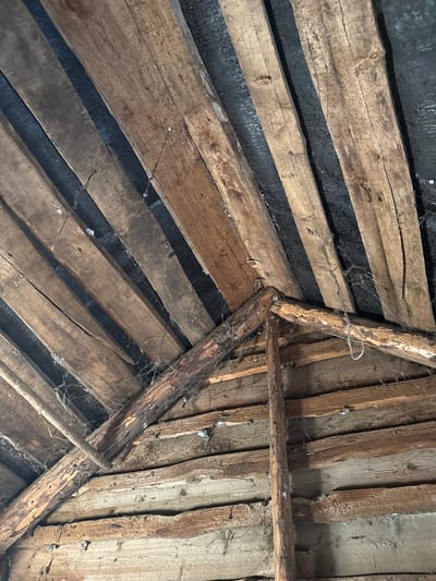 This is a close-up, low-angle photograph of the wooden interior of a rustic structure, possibly a barn or shed, located in Чуй-Атасево, Russia. The scene is dominated by rough-hewn wooden planks, oriented diagonally and horizontally, forming a roof structure. Some planks are weathered and cracked, revealing their age and exposure to the elements. Dark, possibly tarred, material is visible between some of the diagonal planks, suggesting a weatherproofing layer. Cobwebs are present, clinging to the wooden beams and rafters, indicating a lack of recent human activity. No people or animals are visible. The lighting is dim, and the overall impression is one of disuse and age.