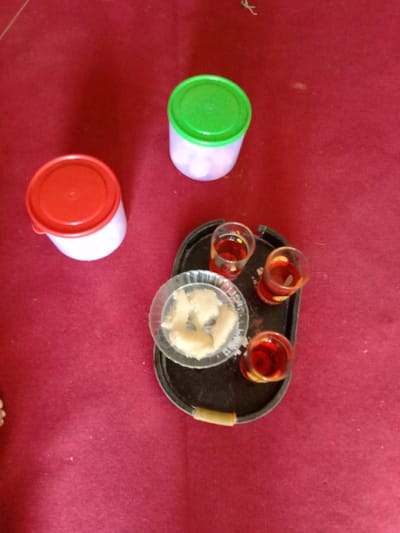 The image presents a still life scene on a dark red surface, likely a tablecloth. There are two circular plastic containers, one red-lidded and one green-lidded, positioned on the left and top of the frame, respectively. A dark-colored tray is placed centrally, containing three small glasses filled with a red liquid, presumably a beverage, and a glass dish holding light-colored, solid pieces, possibly food. The image appears to have been taken indoors. There is no information to confirm that this is in Blangkarieng, Indonesia.