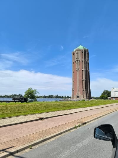 The image shows a tall, brick water tower with a green, conical roof situated next to a body of water in Aalsmeer, Netherlands. The tower is rectangular in shape and has a distinct architectural design with recessed window-like features and vertical brickwork patterns. The water tower is located on a grassy embankment overlooking the water. In the foreground, a paved path runs alongside the road. A black car's side mirror is visible in the lower right corner of the frame. On the grassy area near the water, there is a park bench with a bicycle parked next to it, and a small tree. The sky is blue with some scattered white clouds. A building with a white facade and a dark roof is visible to the right of the water tower.