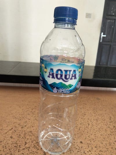 A clear plastic bottle of AQUA mineral water sits upright on a brown textured surface, possibly a table. The bottle is partially filled with water and has a blue screw-on cap. A blue and white label encircles the middle of the bottle, featuring the AQUA logo, an image of lanterns, mountains, and the text "100% MURNI". The background consists of a lightly colored wall and a dark door with a white handle. The scene appears to be indoors, possibly in a home or public establishment. There are no people visible in the image. The image is taken from a slightly low angle, looking up at the bottle. The lighting suggests it is daytime.
