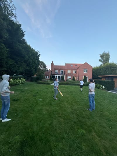 This image depicts an informal cricket game occurring on a large, well-maintained residential lawn within the city of Bubwith, United Kingdom.
The scene features four male individuals. In the left foreground, a male in a light grey hooded sweatshirt and blue jeans holds a small, light-colored ball, appearing ready to bowl. Centered in the mid-ground, a male, possibly a youth, dressed in a light shirt and grey trousers, holds a yellow and black cricket bat in a batting stance. To the right of center in the mid-ground, another male, wearing a white t-shirt and dark shorts/trousers, is captured in motion, running across the grass from right to left. In the right foreground, a fourth male in a light t-shirt and blue jeans stands with his back partially to the viewer, observing the game.
The environment includes an expansive green lawn. In the background, a large, two-story, red-brick house with multiple white-framed windows and a red-tiled roof is visible, featuring a patio or balcony area. Tall, dense green trees and hedges border the property on the left and rear. A small white-framed soccer goal is partially visible in the far left background of the lawn. A smaller, modern-styled outbuilding is present on the far right. The sky is clear and light blue, indicating daylight conditions. Scattered small white debris pieces are visible on the grass in the foreground.