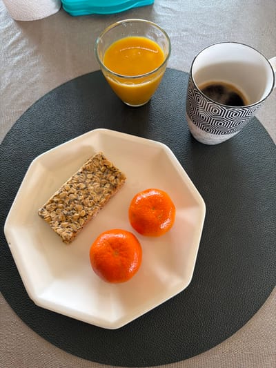 The image displays a breakfast or light meal arrangement, situated in Mauléon-d'Armagnac, France. In the foreground, a light cream-colored, octagonal ceramic plate holds a rectangular granola bar composed of oats and various seeds, positioned alongside two unpeeled, bright orange tangerines. To the upper right of the plate, a white ceramic mug with a repeating black hexagonal geometric pattern contains a dark liquid, consistent with black coffee. To the upper left, a clear glass with a subtle etched pattern on its lower half holds an opaque orange liquid, indicative of fruit juice or a smoothie. All items are arranged on a dark grey or black circular placemat, which rests upon a light grey or beige textured tablecloth or surface. A partially visible roll of white paper towels and a teal plastic container are present in the upper left background.