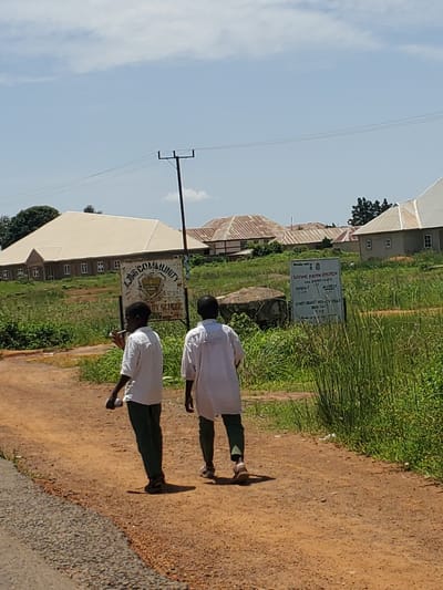 This media file depicts a scene in Mangu, Nigeria, under a clear blue sky with scattered clouds, suggesting it is daytime. Two individuals, likely students given their white uniforms, are walking away from the camera on a dirt path. The path is bordered by a grassy area on the left and a darker, gravelly surface on the right. In the background, several buildings with corrugated iron roofs are visible, indicative of a rural or semi-urban setting.
A signpost stands prominently in the middle ground, displaying text that includes "LIVING FAITH CHURCH," "AKAWATCHA," "MATEK," and "COMMUNITY RY SCHOOL." It also lists service times for "SUNDAY" and "MON SAT." Another signpost is partially visible to the left, with "COMMUNITY" clearly legible. The presence of these signs suggests a community area, possibly near a church and a school. The overall impression is one of a typical day in a Nigerian community, with children on their way somewhere and religious and educational institutions present. The shadows cast by the individuals indicate the sun is relatively high in the sky.